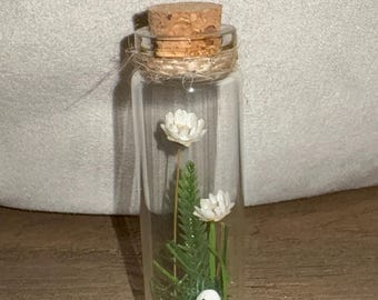 May include: A clear glass bottle with a cork stopper, filled with white flowers, green foliage, and a small white ghost figurine. The bottle is sealed with twine and sits on a wooden surface. A decorative and whimsical item.