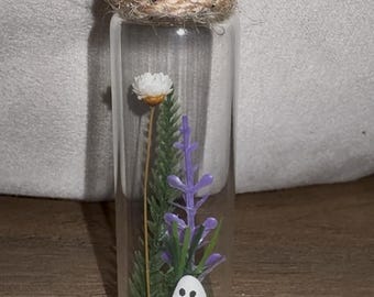 May include: A clear glass bottle with a cork stopper, filled with decorative elements. Inside are a white flower, purple lavender, green foliage, and a small white ghost figure. The bottle is tied with twine.