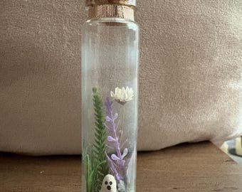 May include: A clear glass bottle with a cork stopper, containing a miniature scene. Inside are sprigs of green and purple plants, a white flower, and a small white ghost figurine. The bottle is decorated with a gold band.