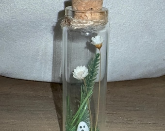 May include: A clear glass bottle with a cork stopper, twine detail, and a miniature ghost figurine inside. The bottle also contains small white flowers and green foliage, creating a whimsical, decorative piece.
