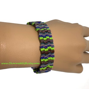 May include: A beaded bracelet featuring a geometric pattern of brown, green, blue, and teal beads. The bracelet is made of small, colorful beads woven together to create a textured surface. The bracelet is shown on a wrist.