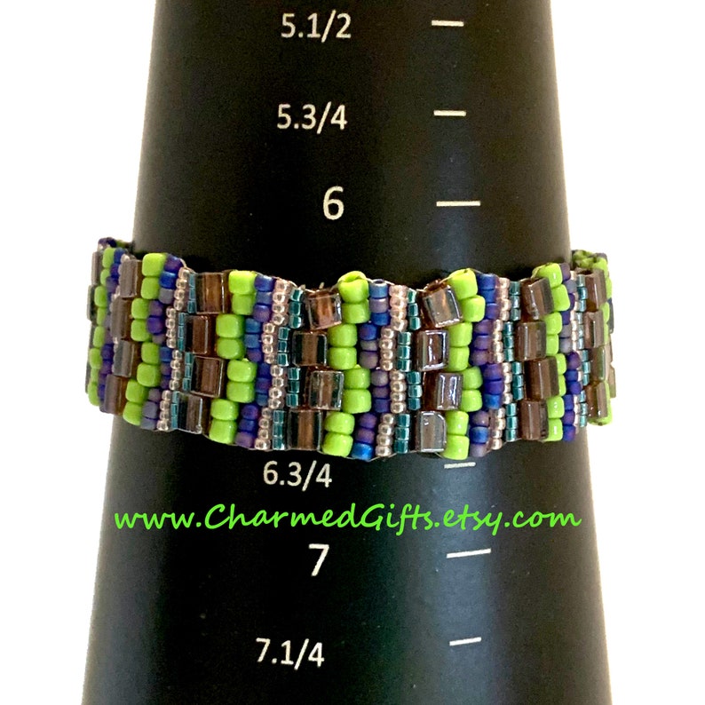 May include: A beaded bracelet in shades of green, blue, and brown is displayed on a black cone-shaped object. The bracelet is made of small, square beads. The text "www.CharmedGifts.etsy.com" is visible.