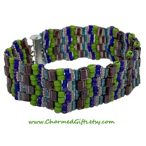 May include: A beaded bracelet featuring a woven pattern of green, blue, and brown beads. The bracelet has a silver-tone clasp and is approximately 1.5 inches wide. The Etsy shop URL www.CharmedGifts.etsy.com is visible at the bottom.