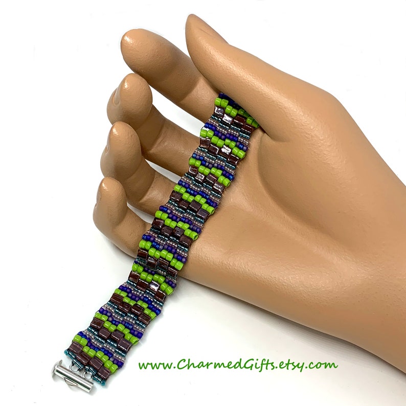 May include: A beaded bracelet with a zig-zag pattern. The bracelet features a combination of green, purple, blue, and brown beads. It has a silver-colored clasp. The website address www.CharmedGifts.etsy.com is visible.