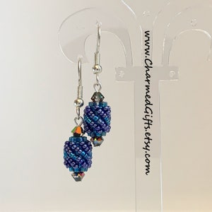 Könnte beinhalten: Ein Paar Hängeohrringe mit silbernen Haken und blauen und türkisfarbenen Perlenkugeln. Die Perlen sind irisierend und haben ein gewebtes Muster.