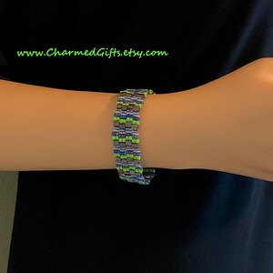 May include: A beaded bracelet in shades of green, blue, gray, and purple. The bracelet is made of small, square beads arranged in a repeating pattern. The text "www.CharmedGifts.etsy.com" is visible in green.