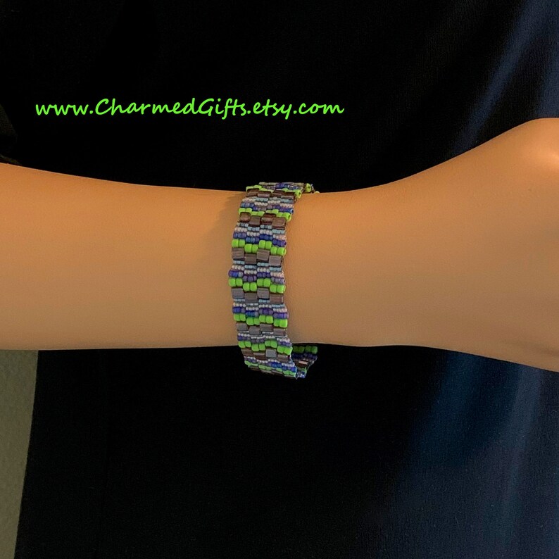 May include: A beaded bracelet in shades of green, blue, gray, and purple. The bracelet is made of small, square beads arranged in a repeating pattern. The text "www.CharmedGifts.etsy.com" is visible in green.