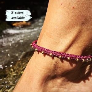 Op de afbeelding: Een enkelbandje van donker magenta gevlochten koord met een reeks kleine, gelijkmatig verdeelde gouden metalen kralen langs de band. De afbeelding bevat een tekst in de linkerbovenhoek met de tekst 8 colors available.