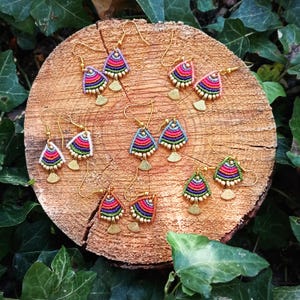 Op de afbeelding: Een collectie van vijf paar waaiervormige oorbellen met kleurrijk geweven draad in roze, paars, groen en blauw. Elke oorbel heeft een goudkleurige metalen haak en een kleine goudkleurige metalen waaiervormige bedel aan de onderkant.