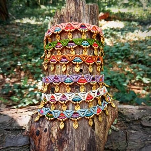 May include: Five stacked macrame bracelets featuring intricate woven patterns in peach, green, orange, purple, white, and blue threads. Each bracelet is accented with small gold-toned metal beads and dangling leaf-shaped charms along the bottom edge.