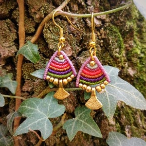 May include: A pair of dangle earrings featuring a triangular macrame design with horizontal stripes in pink, red, purple, and green. Each earring is accented with small gold-toned beads and a textured gold-toned fan-shaped charm at the base.
