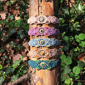 Puede incluir: Cinco pulseras de macramé apiladas verticalmente, cada una con un dije de metal en forma de sol con una piedra preciosa redonda. Los patrones de cordón trenzado son de color rosa claro, morado, azul claro, beige y verde azulado oscuro.