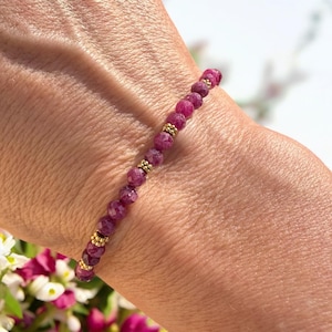 Op de afbeelding: Een delicate armband gemaakt met kleine, gefacetteerde robijn kralen en gouden spacers. De armband wordt getoond op de pols van een persoon.