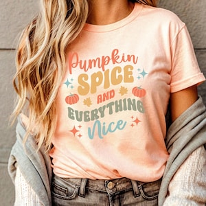 May include: Peach-coloured t-shirt with the text "Pumpkin Spice and Everything Nice" in a retro font. The design includes pumpkins, leaves, and star accents. The shirt is short-sleeved and has a crew neck.
