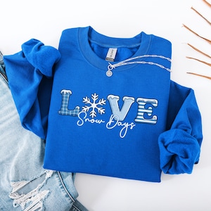Może przedstawiać: Królewska niebieska bluza z napisem "LOVE" w zimowym motywie. Litera "L" ma niebieski wzór w kratę, "O" to płatek śniegu, a "VE" jest niebieskie z białymi akcentami. Słowa "Snow Days" są napisane kursywą poniżej.