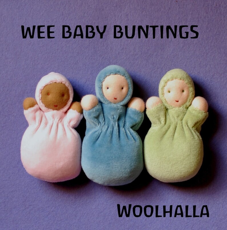 Pu&ograve; includere: Tre bambole Wee Baby Buntings rosa, blu e verdi, con dettagli marroni, bianchi e rosa. Le bambole hanno un viso semplice e sono realizzate in tessuto morbido. Il testo "WEE BABY BUNTINGS" e "WOOLHALLA" sono visualizzati.