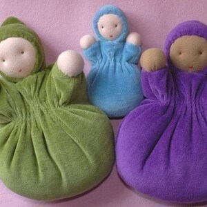 Pu&ograve; includere: Tre bambole di stoffa fatte a mano nei toni del verde, blu e viola. Ogni bambola ha un corpo rotondo, una testa con cappuccio e braccia piccole e arrotondate. I volti sono semplici, con tratti cuciti. Le bambole sono disposte su una superficie rosa.