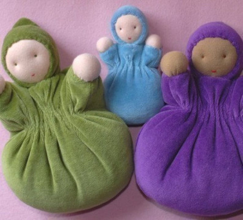 Pu&ograve; includere: Tre bambole di stoffa fatte a mano nei toni del verde, blu e viola. Ogni bambola ha un corpo rotondo, una testa con cappuccio e braccia piccole e arrotondate. I volti sono semplici, con tratti cuciti. Le bambole sono disposte su una superficie rosa.