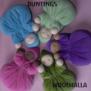 Pu&ograve; includere: Una collezione di bambole per bambini in peluche colorate, in vari colori tra cui blu, verde, viola, rosa e verde. Ogni bambola ha un viso rotondo e un abito con cappuccio. Le parole "BUNTINGS" e "WOOLHALLA" sono visibili.