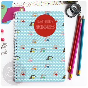 May include: A blue spiral notebook with a pattern of swimmers and waves. The cover has a red circle with the text "LAYERED SWIMMERS" in white.