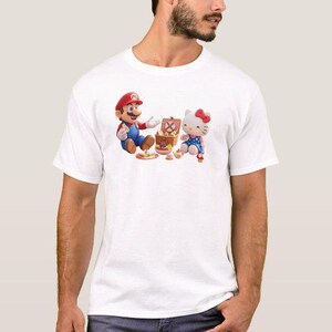 May include: White t-shirt featuring a colorful graphic of Mario and Hello Kitty having a picnic. Mario is wearing his signature red hat and blue overalls. Hello Kitty is in a blue dress with a red bow. A picnic basket and food are also visible.