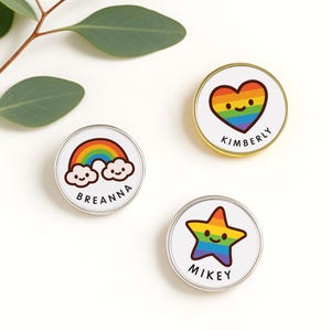 May include: Three circular pins with rainbow designs. One pin shows a rainbow with smiling clouds and the name "Breanna". Another has a rainbow heart with "Kimberly". The third pin has a rainbow star with "Mikey". The pins have silver and gold rims.