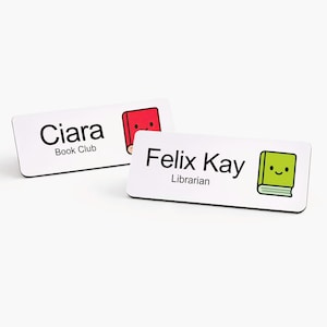 May include: Two white name badges with black text and cartoon book illustrations. One badge reads "Ciara Book Club" with a red book graphic. The other reads "Felix Kay Librarian" with a green book graphic.