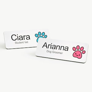 May include: Two white name tags with black text and paw print designs. One tag reads "Ciara, Student Vet" with a blue paw print. The other reads "Arianna, Dog Groomer" with a pink paw print.