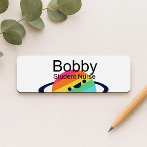 May include: A white name tag with the name "Bobby" in black text. Below the name is the text "Student Nurse". A colorful planet graphic with a smiling face is below the text. A pencil is in the bottom right corner.