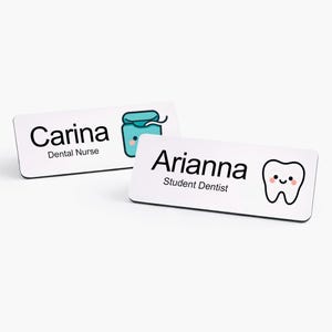 May include: Two white name tags with black borders. One tag reads "Carina, Dental Nurse" with a cartoon floss container. The other tag reads "Arianna, Student Dentist" with a smiling tooth graphic.
