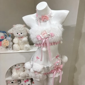 May include: A white faux fur tube top and matching shorts set featuring pink gingham trim, satin ribbons, and small bells. The top includes a halter neck strap with a pink bow, while the shorts are adorned with plush white pom-poms and bows.