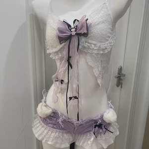 May include: A white lace camisole with a lavender and black ribbon bow featuring a small character charm, paired with lavender denim shorts that have frayed edges, white ruffled lace trim, and two small white plush ears on the waistband.