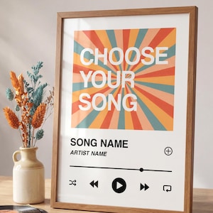 May include: Framed print with the text "CHOOSE YOUR SONG" against a retro-style burst of orange, teal, and peach. Below, a music player interface design is displayed. The frame is a light brown color.