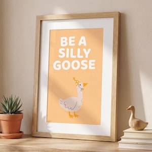 May include: Framed art print with the words "BE A SILLY GOOSE" in white block letters on an orange background. A cartoon goose wearing a floral crown is below the text. The frame is light brown.