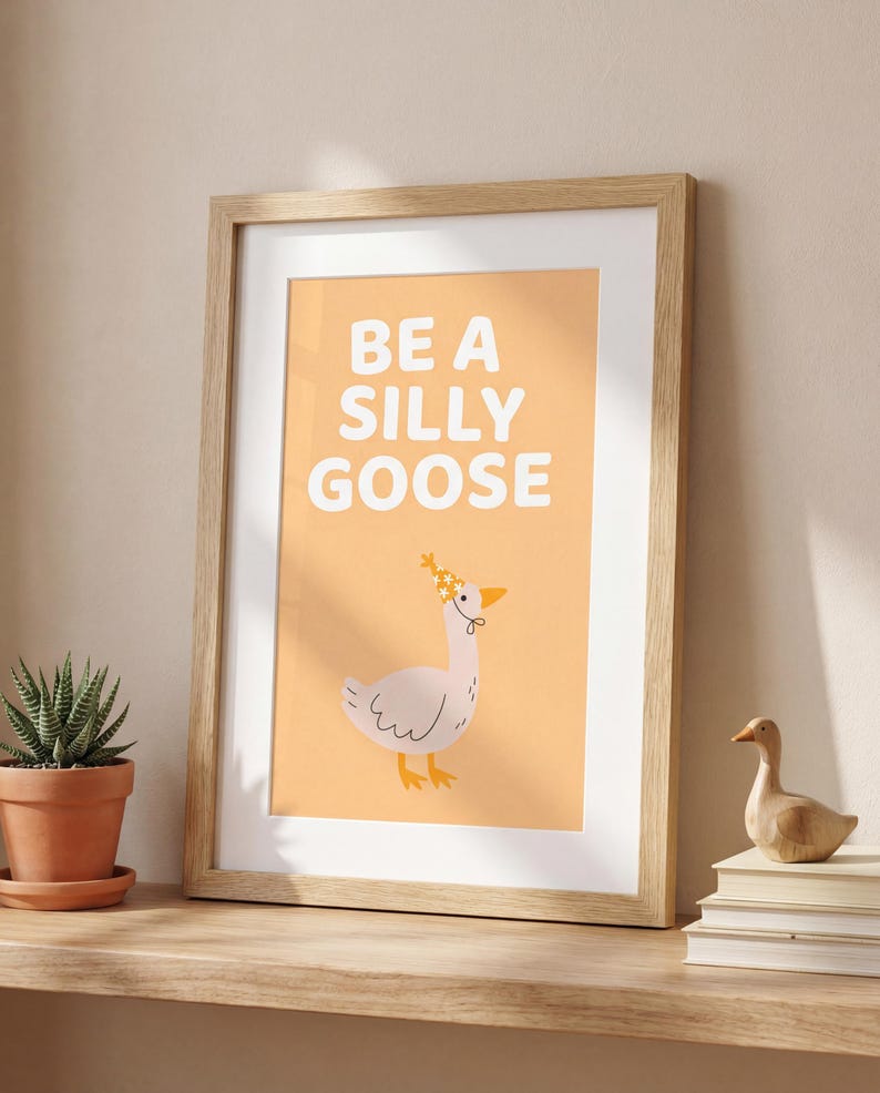 May include: Framed art print with the words "BE A SILLY GOOSE" in white block letters on an orange background. A cartoon goose wearing a floral crown is below the text. The frame is light brown.