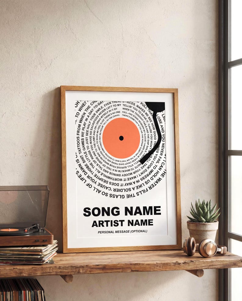 May include: A framed print featuring a vinyl record design with a circular arrangement of text. The record's center is orange, and the text includes the words "Song Name" and "Artist Name" in bold, black font. The print is displayed on a wooden shelf.