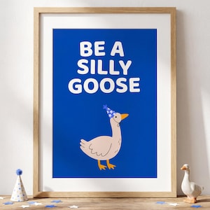 May include: A framed art print with the words "BE A SILLY GOOSE" in white on a blue background. The print features a cartoon goose wearing a party hat. The artwork is displayed on a wooden surface with decorative items.