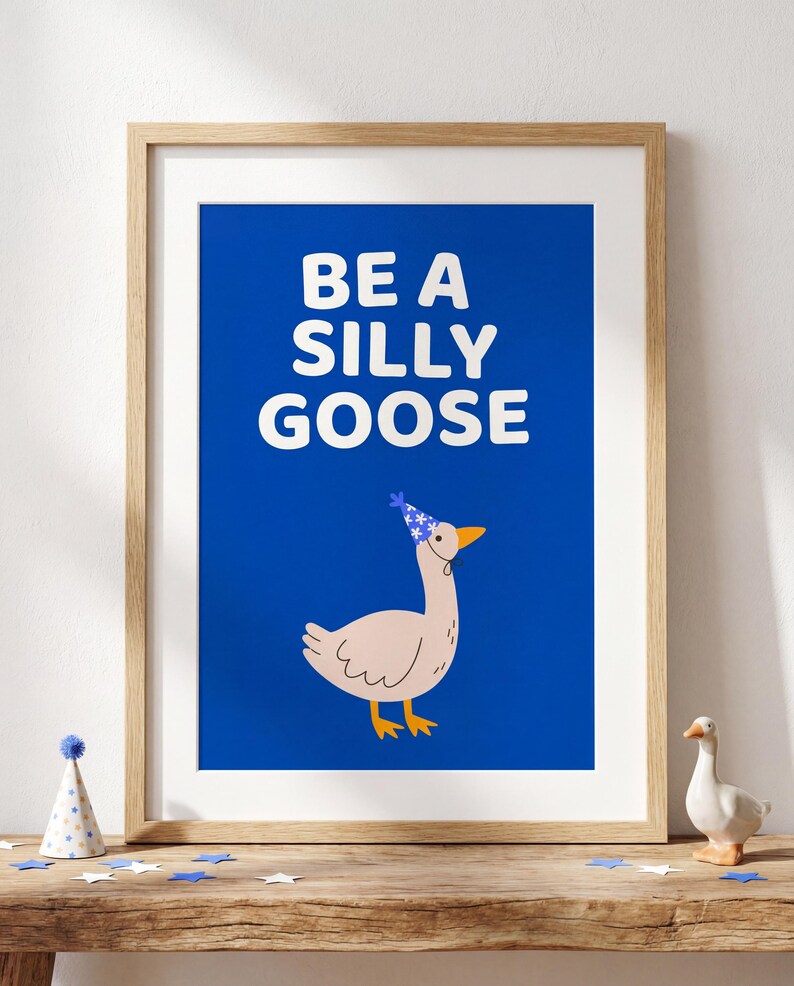 May include: A framed art print with the words "BE A SILLY GOOSE" in white on a blue background. The print features a cartoon goose wearing a party hat. The artwork is displayed on a wooden surface with decorative items.