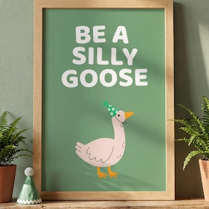 May include: A framed art print with the text "BE A SILLY GOOSE" on a green background. The print features a cartoon goose wearing a party hat. The frame is made of light-colored wood. Ferns in terracotta pots are on either side of the print.