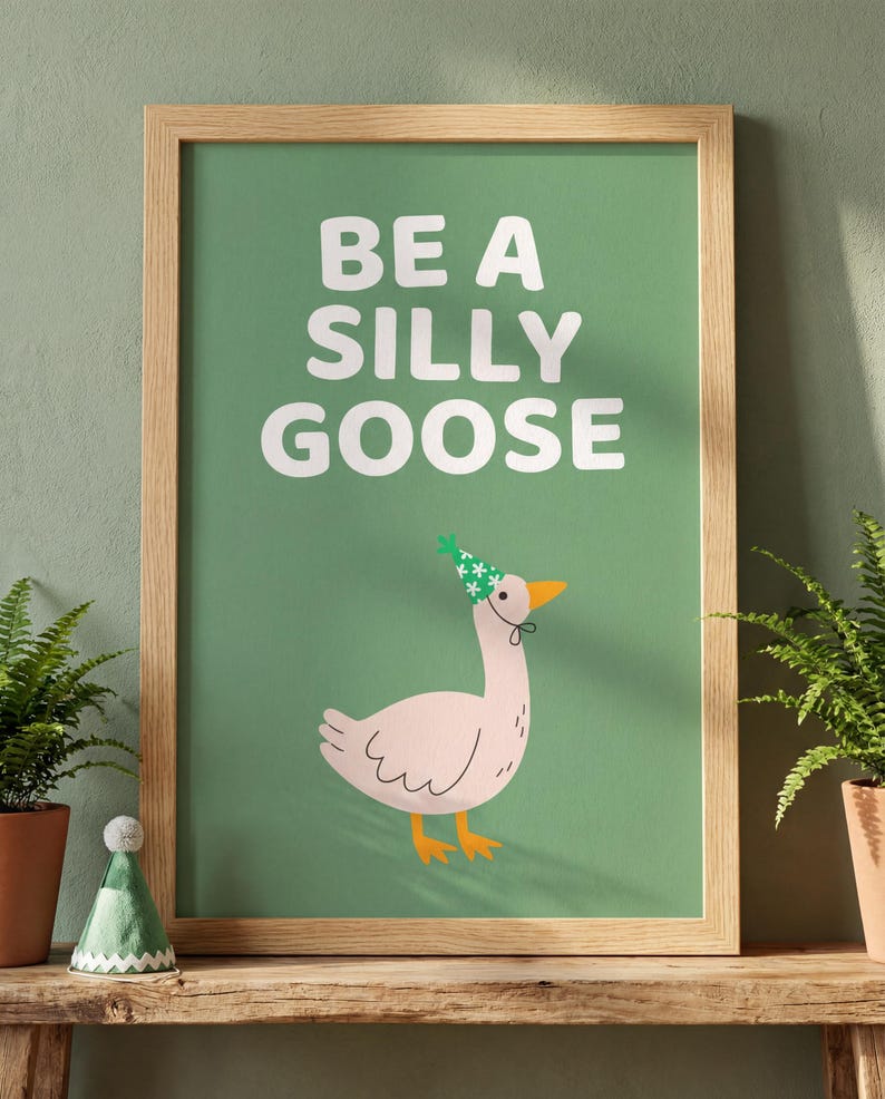 May include: A framed art print with the text "BE A SILLY GOOSE" on a green background. The print features a cartoon goose wearing a party hat. The frame is made of light-colored wood. Ferns in terracotta pots are on either side of the print.