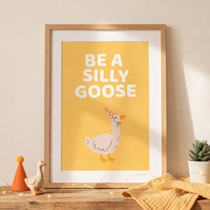 May include: A framed art print with the words "BE A SILLY GOOSE" in white on a yellow background. The print features a cartoon goose wearing a party hat. A wooden shelf holds the framed print, a small goose figurine, and a potted succulent.
