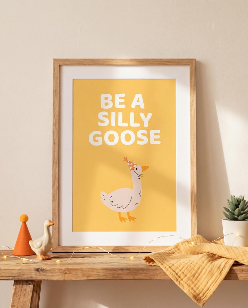 May include: A framed art print with the words "BE A SILLY GOOSE" in white on a yellow background. The print features a cartoon goose wearing a party hat. A wooden shelf holds the framed print, a small goose figurine, and a potted succulent.
