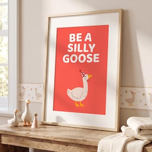 May include: Framed art print with a red background and the words "BE A SILLY GOOSE" in white. The print features a cartoon goose wearing a party hat. The artwork is displayed on a wooden table with decorative geese figurines.