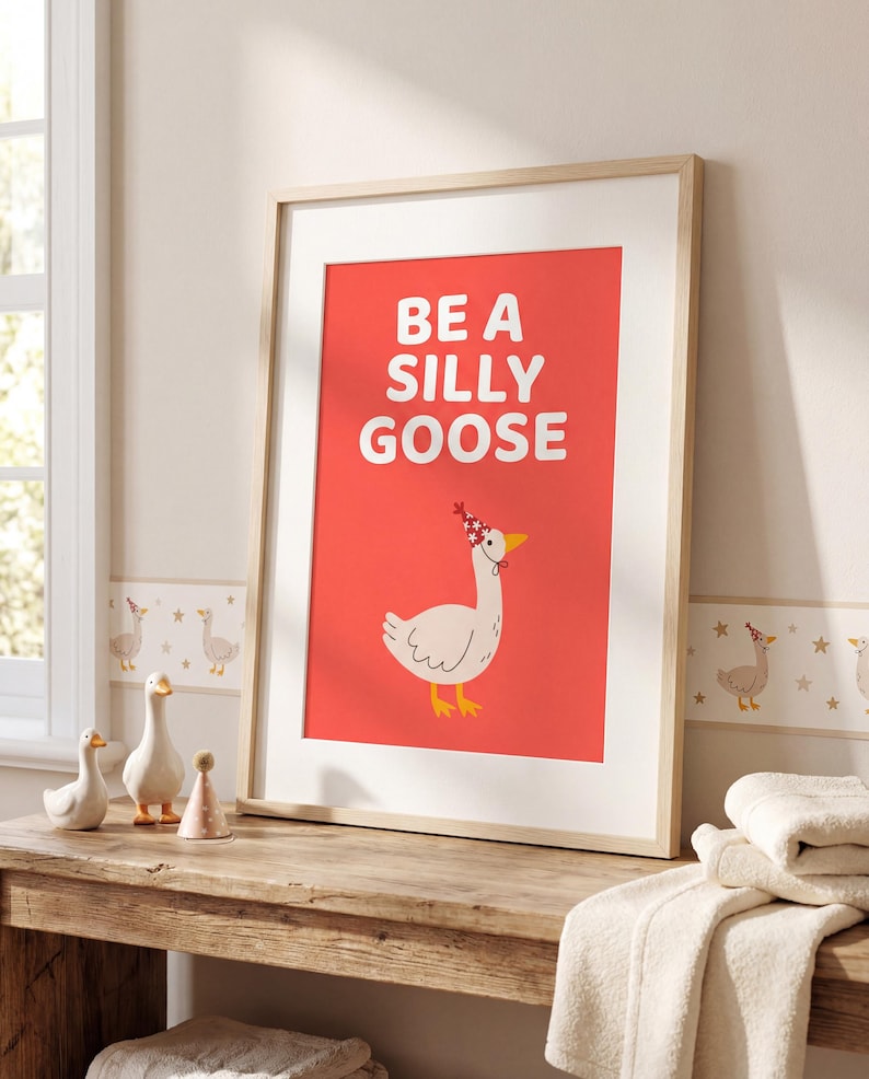 May include: Framed art print with a red background and the words "BE A SILLY GOOSE" in white. The print features a cartoon goose wearing a party hat. The artwork is displayed on a wooden table with decorative geese figurines.