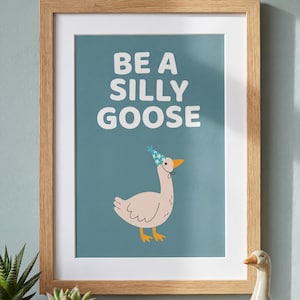 May include: A framed art print with the words "BE A SILLY GOOSE" in white block letters. The print features a cartoon goose wearing a party hat. The frame is light brown.