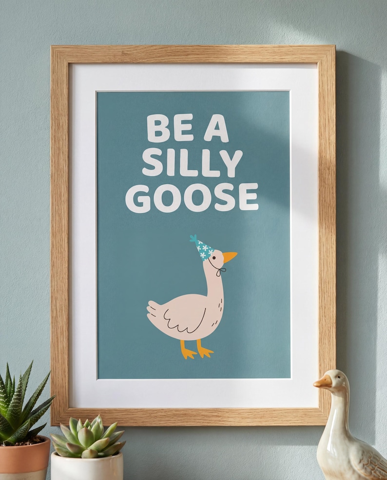 May include: A framed art print with the words "BE A SILLY GOOSE" in white block letters. The print features a cartoon goose wearing a party hat. The frame is light brown.