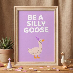 May include: A framed print with the text "BE A SILLY GOOSE" on a purple background. The print features a cartoon goose wearing a party hat. A small ceramic goose figurine and confetti are on a wooden surface.