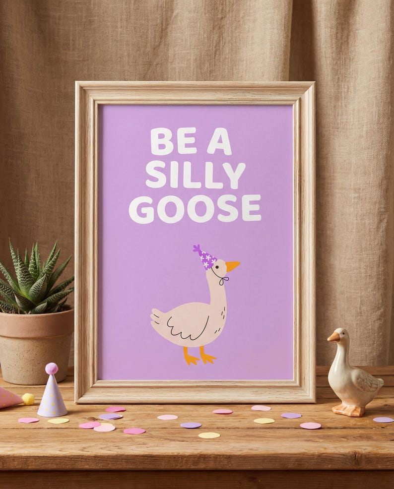 May include: A framed print with the text "BE A SILLY GOOSE" on a purple background. The print features a cartoon goose wearing a party hat. A small ceramic goose figurine and confetti are on a wooden surface.