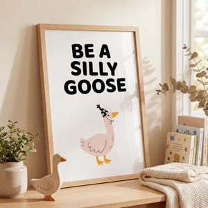 May include: A framed print with the words "BE A SILLY GOOSE" in black letters. Below the text is an illustration of a pink goose wearing a party hat. A wooden goose figurine sits on a wooden table.