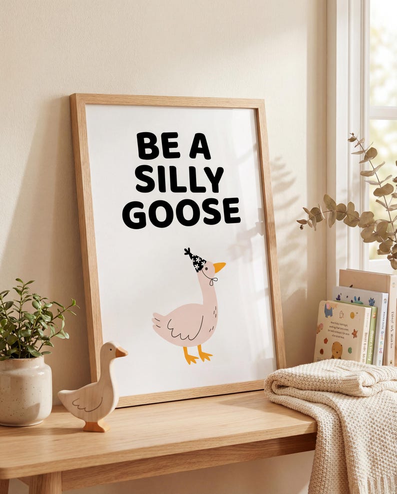 May include: A framed print with the words "BE A SILLY GOOSE" in black letters. Below the text is an illustration of a pink goose wearing a party hat. A wooden goose figurine sits on a wooden table.